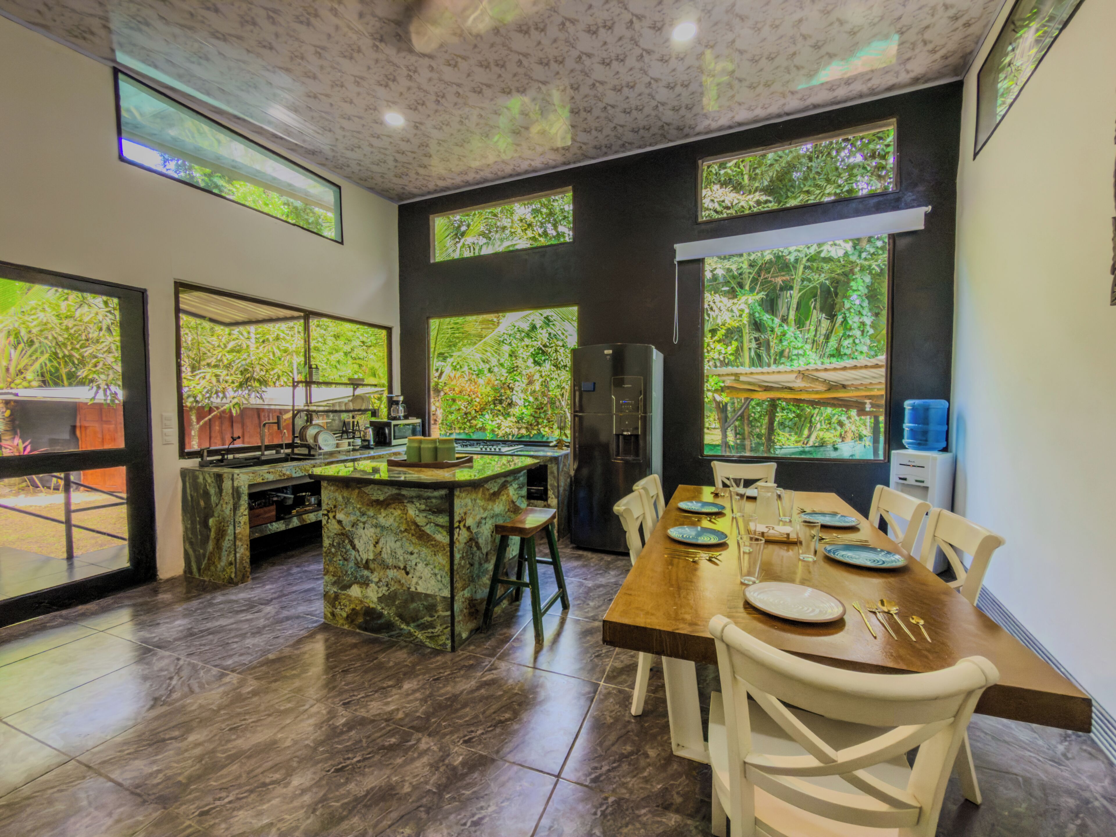 Une cuisine moderne dotée d’un îlot en marbre, de grandes fenêtres et d’un coin repas avec des chaises et une table dressée pour quatre personnes.