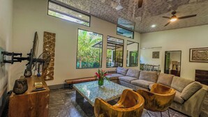 A spacious living area with a sofa, wooden chairs, a coffee table, and a fan. There are large windows providing natural light and views of greenery outside. - Mountain House,Beautiful 3-bedroom with AC in quaint Puerto Viejo de Talamanca (Puerto Viejo de Talamanca)