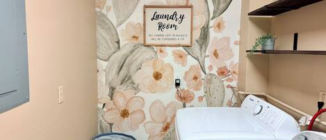 A laundry room with a floral wallpaper, a framed sign, a washing machine, and a blue laundry basket.