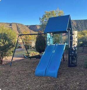 Ein Spielplatz mit Rutsche und Schaukeln, umgeben von Bäumen und Hügeln.