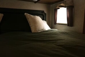 A bed with a dark headboard and a single white pillow, a window with curtains, and a wooden ceiling. - Forest Glamping Getaway with King Bed, Deck, and Cozy Fireplace (Dallas)