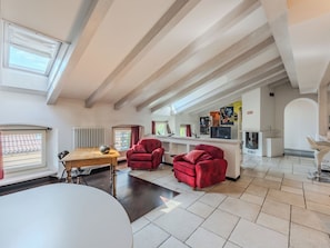 A spacious living area with a high ceiling, a dining table, red armchairs, and a kitchen area with a TV. - Le Falesie by Interhome (Arco)
