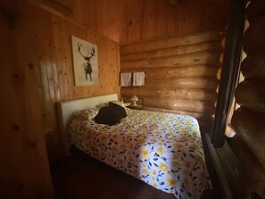 A wooden cabin room with a bed, a wall-mounted painting of a deer, and a window with curtains. - The Big Log Cabin (Saint-Gabriel-de-Valcartier)