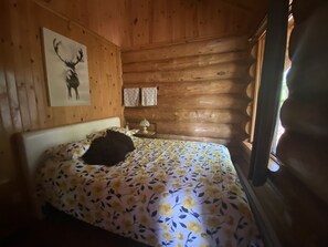 A wooden cabin room with a bed, a wall-mounted picture, and a window. - The Big Pine Shelter (Saint-Gabriel-de-Valcartier)