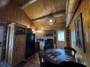 A wooden interior with a dining table, chairs, a refrigerator, and a kitchen area with a stove and sink. - The Big Pine Shelter (Saint-Gabriel-de-Valcartier)