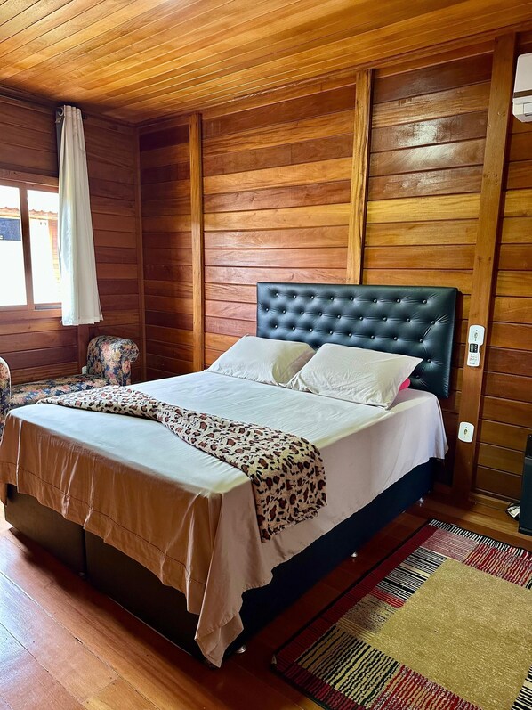 A wooden-paneled room with a bed, a window with a curtain, and a patterned rug.