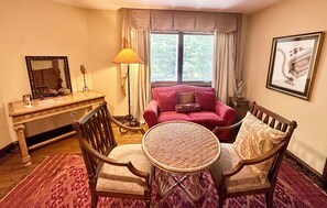 Ein Zimmer mit einem roten Sofa, einem runden Korbstuhl, einem Holzstuhl, einem Schreibtisch mit Lampe und einem Fenster mit Vorhängen.