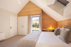 A bedroom with a sloped ceiling, a bed with pillows, a nightstand with a lamp, and a window with curtains. - Speargrass Villa | Luxury | Hot Tub | Pool (Queenstown)