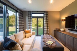 A modern living room with a sofa, ottoman, and a flat-screen TV mounted on the wall. - Speargrass Villa | Luxury | Hot Tub | Pool (Queenstown)