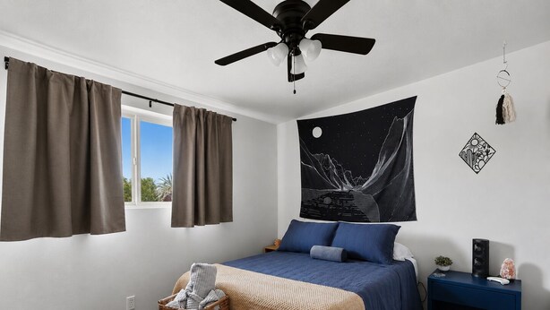 Un dormitorio con cama, ventilador de techo, tapizris de cielo nocturno, mesita de noche pequeña y ventana con cortinas.