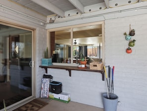 A room with a glass door, a window with a view, a shelf with plants, a ladder toss game box, and a trash can. - New Relaxing 3 Bedroom Desert Oasis with Sparkling Pool! (Tucson)