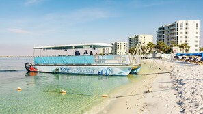 A beachside boat with a covered deck and passengers, a sandy beach with umbrellas, and a hotel in the background. - Beautiful condo with amazing view located in Emerald Grande at the Harborwalk. (Destin)