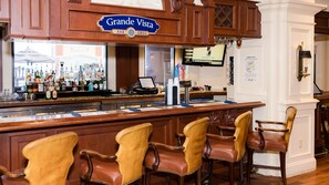A bar area with a sign reading 'Grande Vista BAR GRILL,' bar stools, a counter with various bottles, and a television screen. - Beautiful condo with amazing view located in Emerald Grande at the Harborwalk. (Destin)