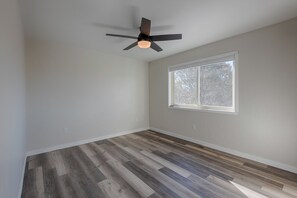 Une pièce dotée d’un ventilateur de plafond, de planchers en bois, d’une grande fenêtre donnant sur la verdure et d’un mur blanc.