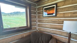 A cozy room with a wooden log wall, a window with a view of a green landscape, a framed picture, and a small table with a lamp.