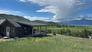 A rustic cabin with a porch, surrounded by a grassy field and mountains in the background.