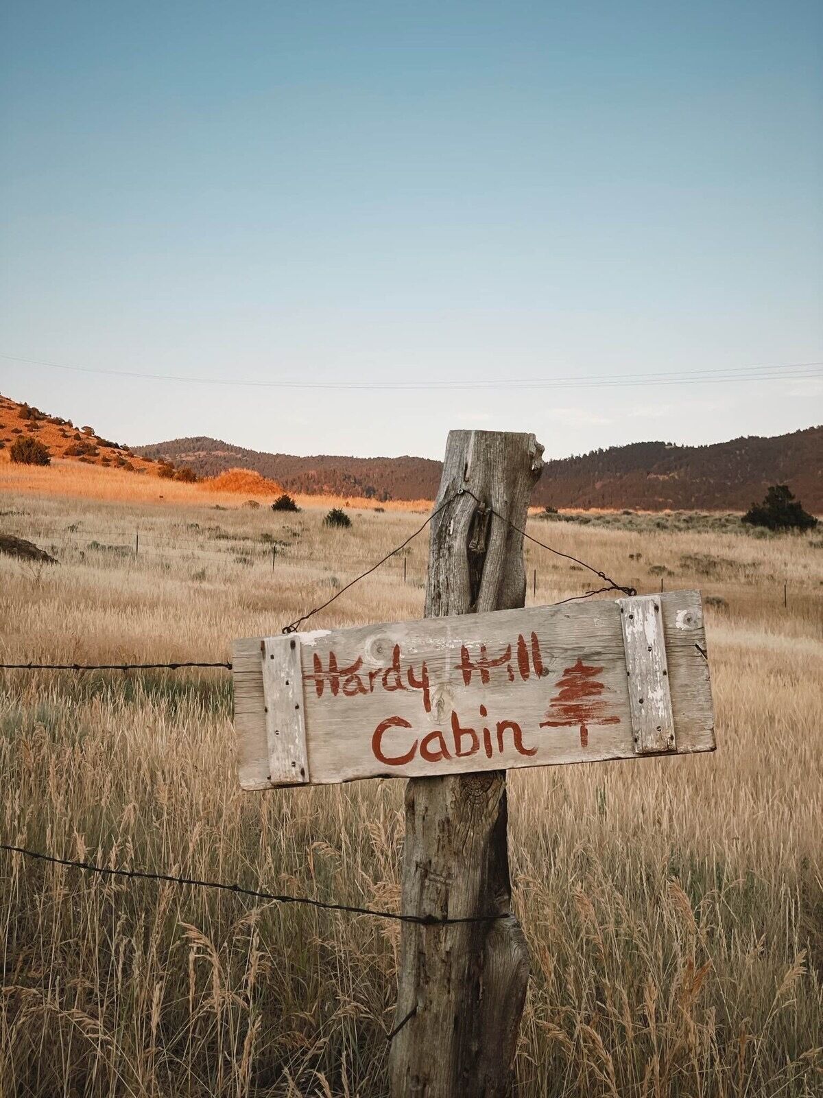 A wooden signpost with 'Hardy Hill Cabin' written on it, set against a backdrop of a grassy field and hills.
