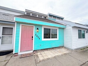 Uma casa de dois andares com fachada azul-turquesa, uma porta rosa e o número '5628' na lateral.