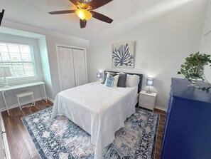 A bedroom with a bed, bedside tables, a desk, a ceiling fan, a window with blinds, and a decorative wall piece. - Beach Breeze - Across from Beach (Panama City)