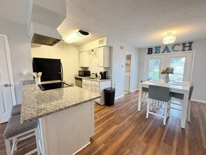 A kitchen with granite countertops, a black refrigerator, and a microwave. There is a trash can and a dining area with a table and chairs. The word 'BEACH' is visible on the wall. - Beach Breeze - Across from Beach (Panama City)