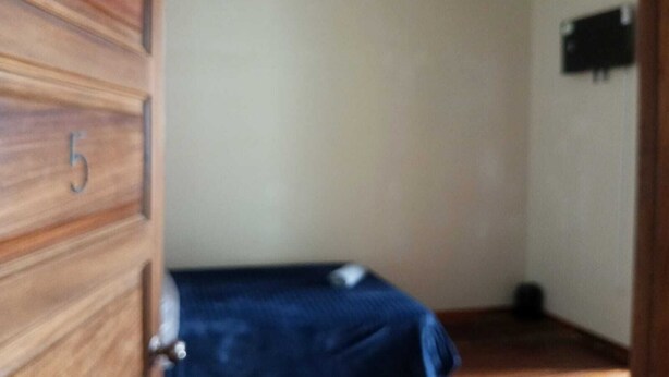 Una habitación con una puerta de madera marcada con el número 5, una cama con cubrecolcha azul y un pequeño objeto negro en la pared.