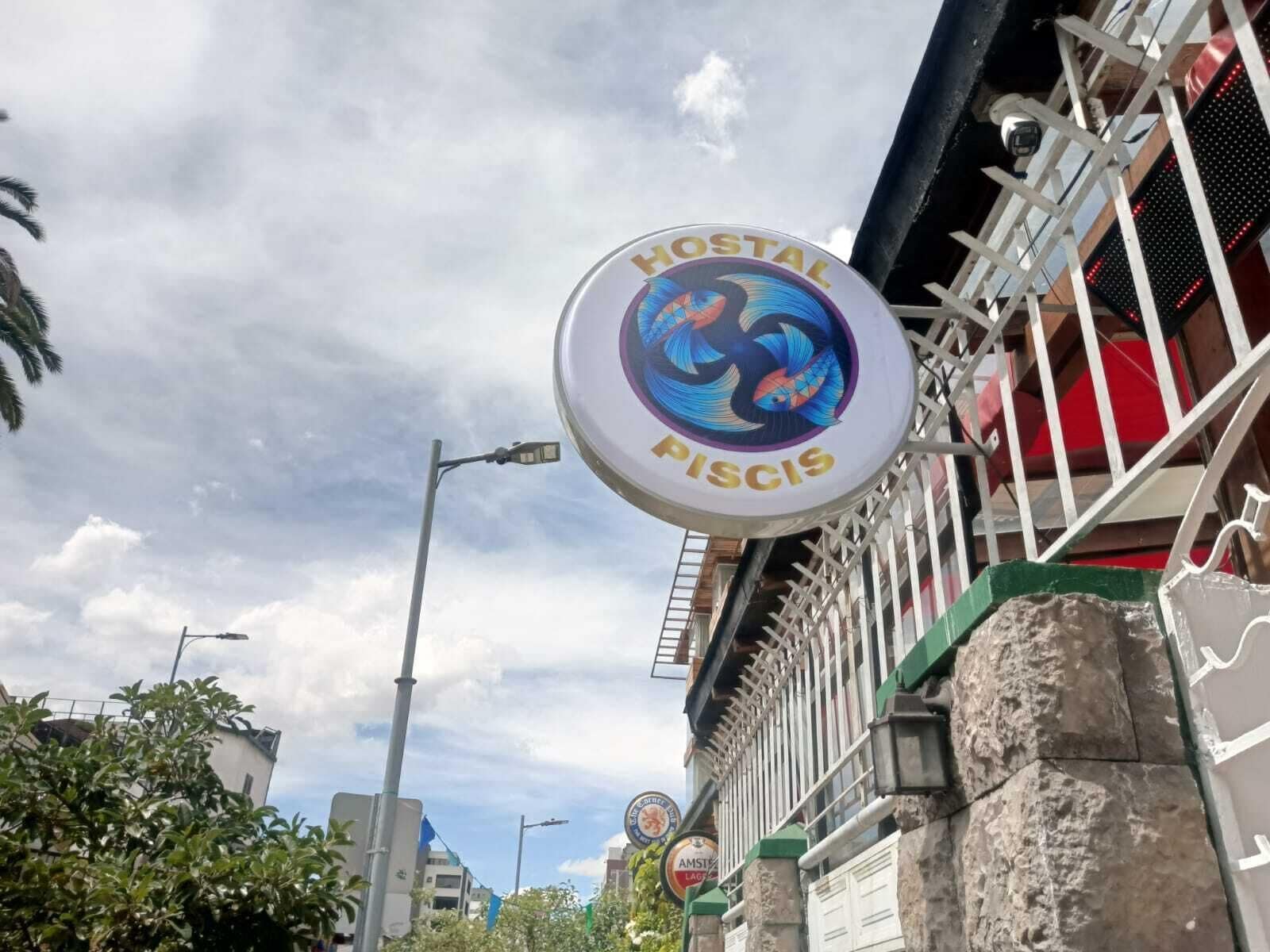 A hostel sign with a colorful logo and the name 'HOSTAL PISCIS' against a partly cloudy sky.