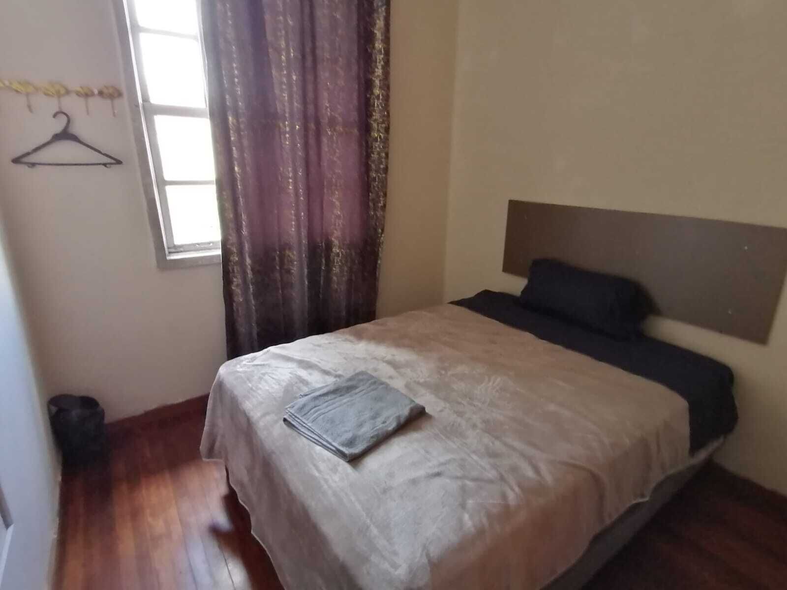 A bedroom with a bed, a window with curtains, a hanging coat rack, and a trash bin.