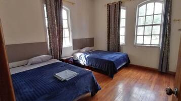 A room with two beds, wooden flooring, and large windows with curtains.