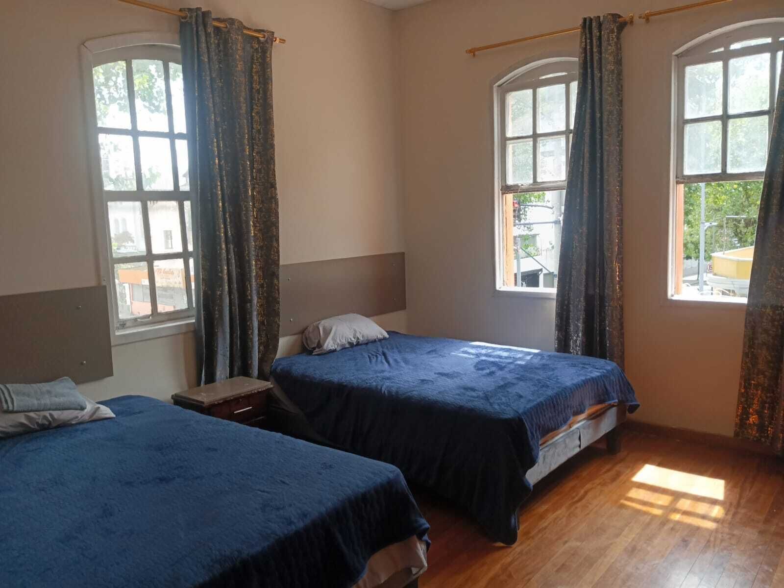 Una habitación con dos camas, piso de madera y ventanas amplias con cortinas.