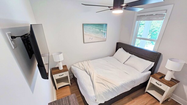 Quarto com cama, mesas de cabeceira, ventoinha no teto, janela e um quadro na parede.