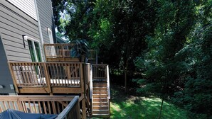 Um deck de madeira leva a uma casa com área coberta, cercada por um gramado e árvores.