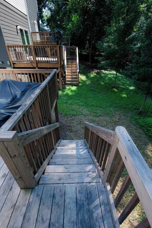 Eine Holzterrasse führt zu einem Haus mit überdachtem Bereich, umgeben von einem Rasen und Bäumen.