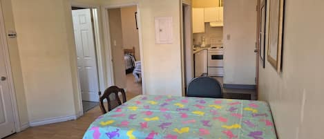 A dining area with a table covered by a colorful tablecloth, wooden chairs, and a kitchen in the background.