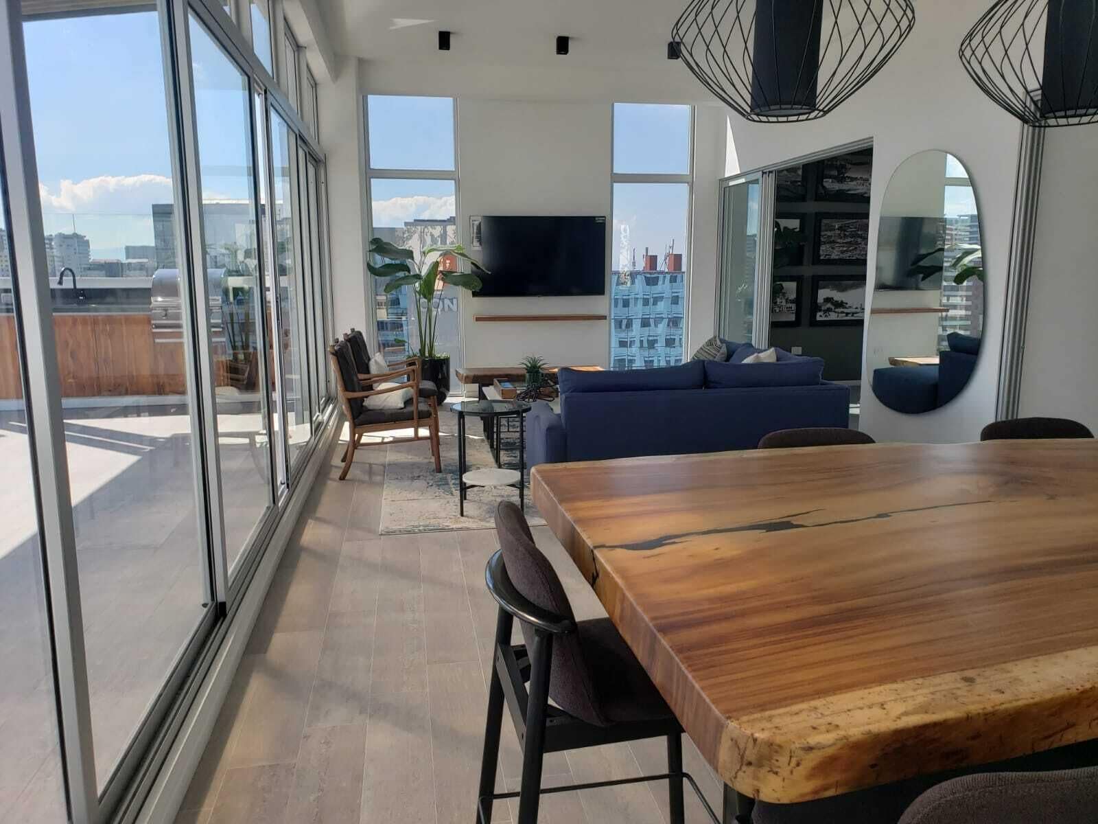 A modern living space with a dining area, a sofa, a TV, and large windows offering a city view.