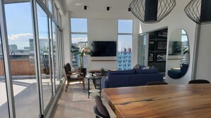 Un espacio de estar moderno con comedor, sofá, televisor y ventanas amplias con vista a la ciudad.