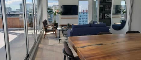 A modern living space with a dining area, a sofa, a TV, and large windows offering a city view.