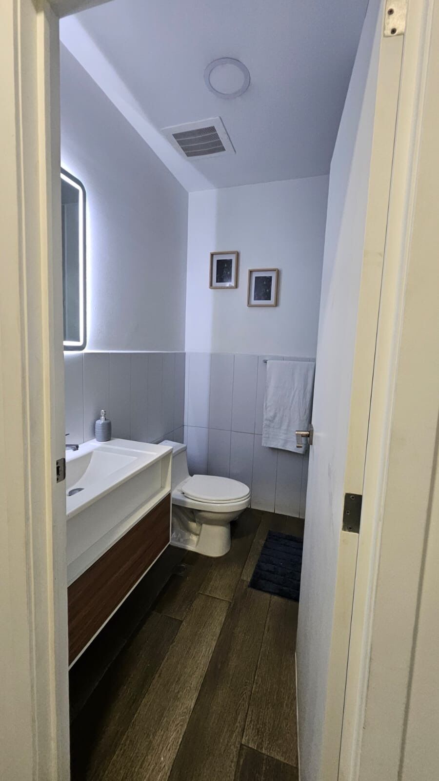Une petite salle de bain avec un lavabo blanc, des toilettes et un miroir. Deux tableaux encadrés sont accrochés au mur, et une serviette est suspendue au mur.
