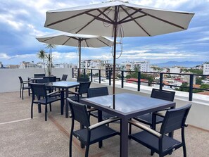 An outdoor terrace with tables and chairs, umbrellas, and a view of the cityscape. - Serenity Tower 601, 1-Bedroom Apartment with Rooftop Pool and Ocean and Mountain Views (Puerto Vallarta)