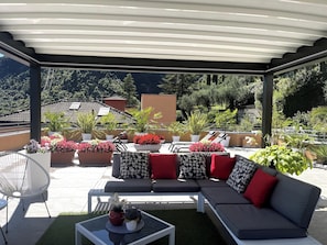 A covered outdoor seating area with a sofa, chairs, and potted plants. - Apartment with a 150-square-meter Private Terrace and Breathtaking Lake Views (Trentino-Alto Adige)