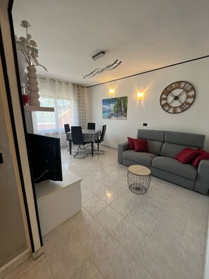 A living room with a grey sofa, a dining table with chairs, a TV, and a painting on the wall. - Apartment with a 150-square-meter Private Terrace and Breathtaking Lake Views (Trentino-Alto Adige)