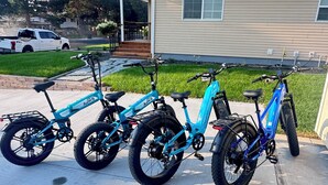 Fyra blå elcyklar står parkerade på en betonguppfart framför ett hus med ett tak av metall.