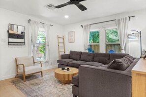 A modern living room with a grey sectional sofa, a wooden coffee table, and a chair. There is a ladder leaning against the wall, a ceiling fan, and a window with curtains. - Chef`s Kitchen + Fire Pit Patio | E Austin Bungalow Mins to Local Favs & Lady Bird (Austin)