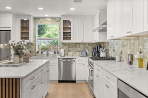 A modern kitchen with white cabinetry, a stainless steel dishwasher, and a large window overlooking a garden.