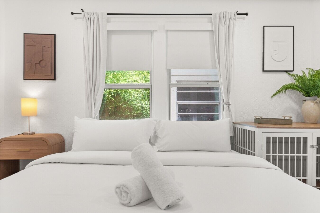 Una cama bien hecha con ropa de cama blanca, una lámpara de noche, una ventana con cortinas, un radiador y un cuadro en la pared.