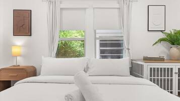A neatly made bed with white bedding, a bedside lamp, a window with curtains, a radiator, and a framed picture on the wall.