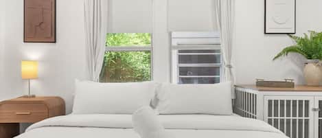 Una cama bien hecha con ropa de cama blanca, una lámpara de noche, una ventana con cortinas, un radiador y un cuadro en la pared.