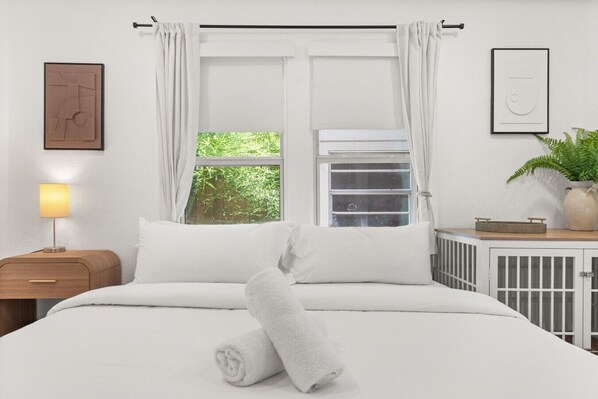 Una cama bien hecha con ropa de cama blanca, una lámpara de noche, una ventana con cortinas, un radiador y un cuadro en la pared.