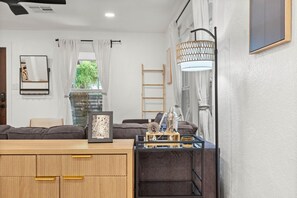 Una habitación con un escritorio de madera, un cuadro enmarcado, una lámpara, una escalera y una ventana con cortinas.
