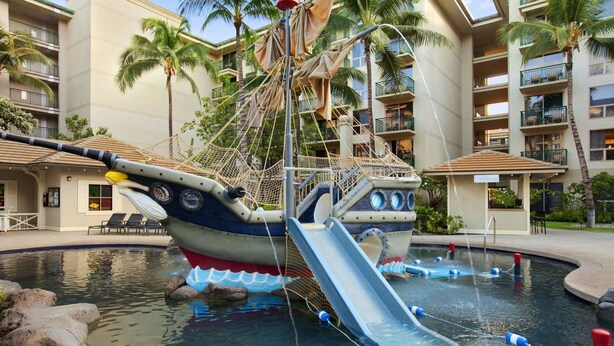 Zona de piscina con estructura de juego temática pirata, que incluye un tobogán y una zona de escalada con red.