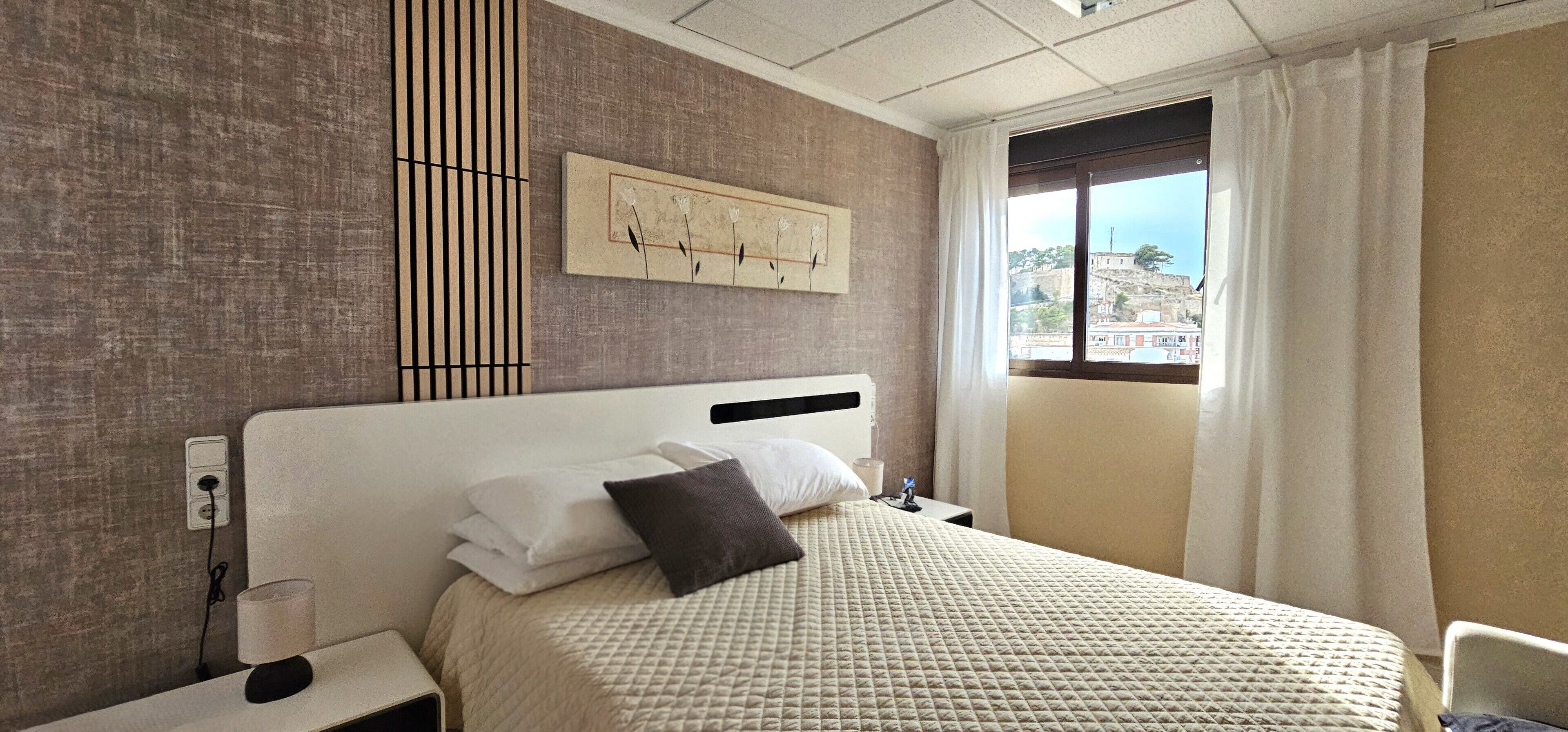 A hotel room with a bed, a nightstand, a power outlet, a window with curtains, and a wall-mounted artwork.
